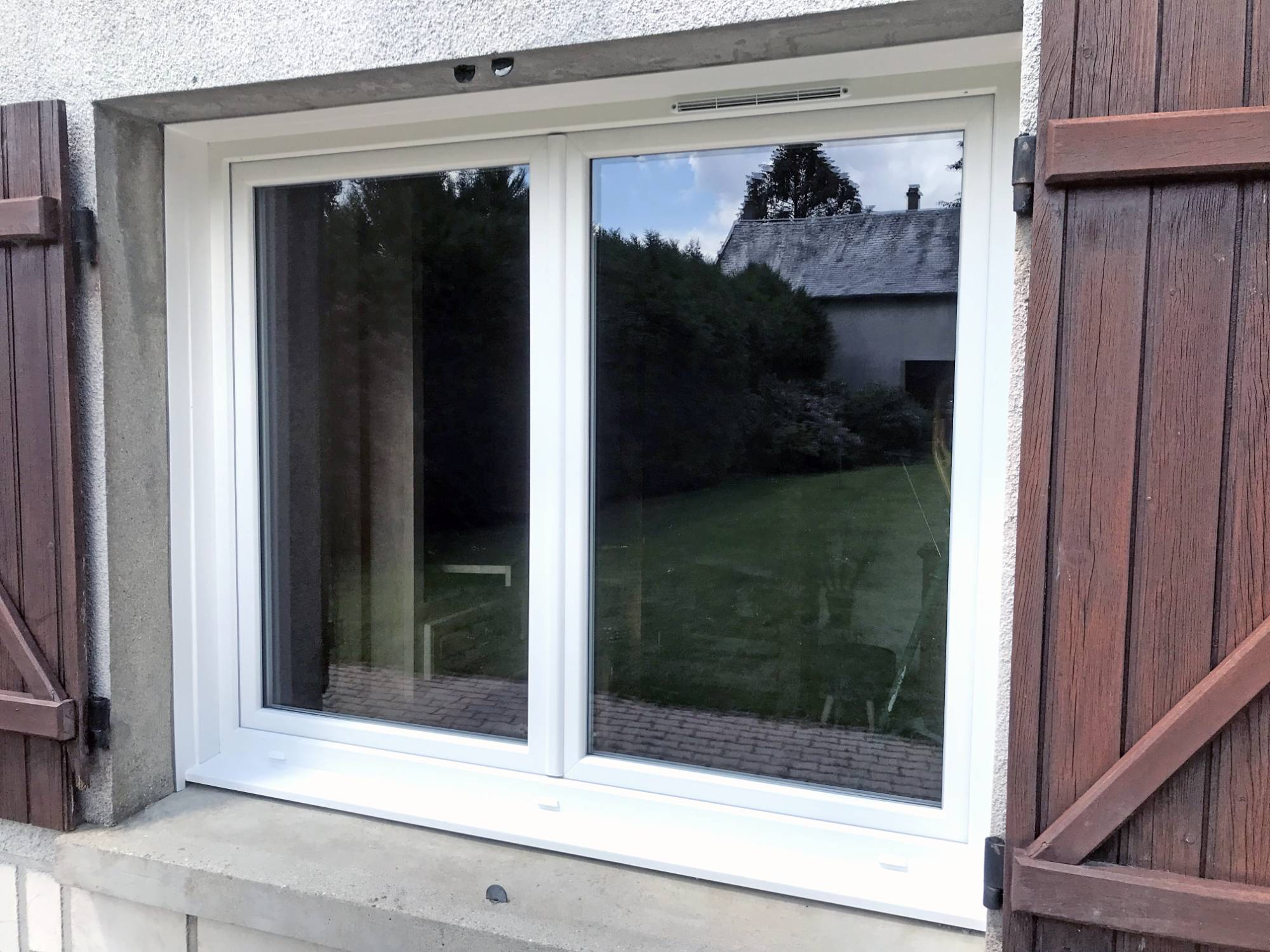 Fenêtre en PVC avec volets en bois, donnant sur un jardin verdoyant.