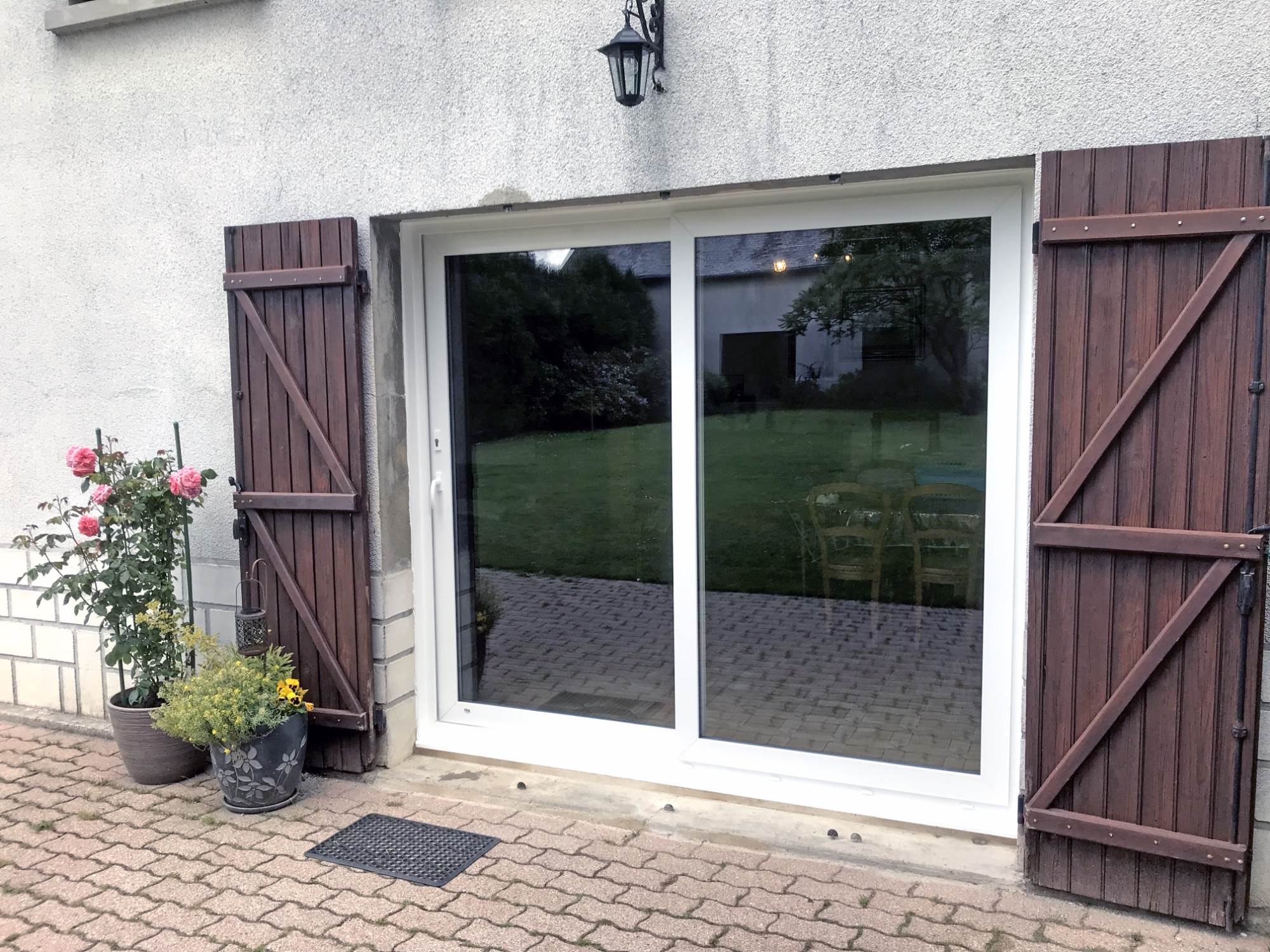 Porte-fenêtre avec volets, fleur en pot et vue sur le jardin.