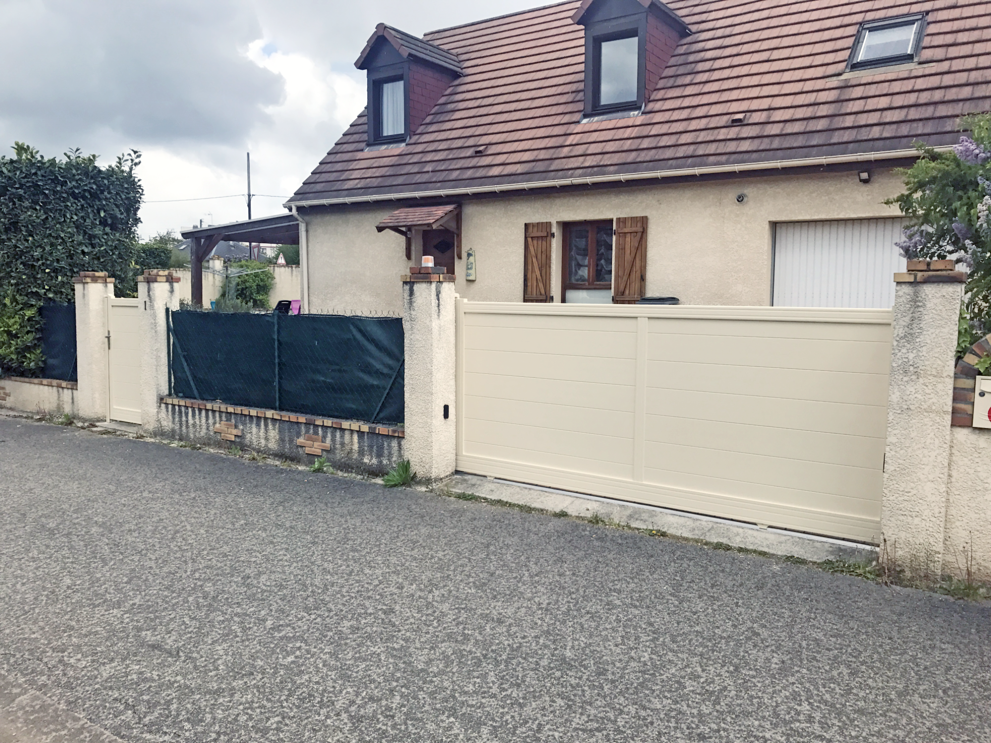 Maison avec toit en pente, clôture beige et jardin, ciel nuageux en arrière-plan.