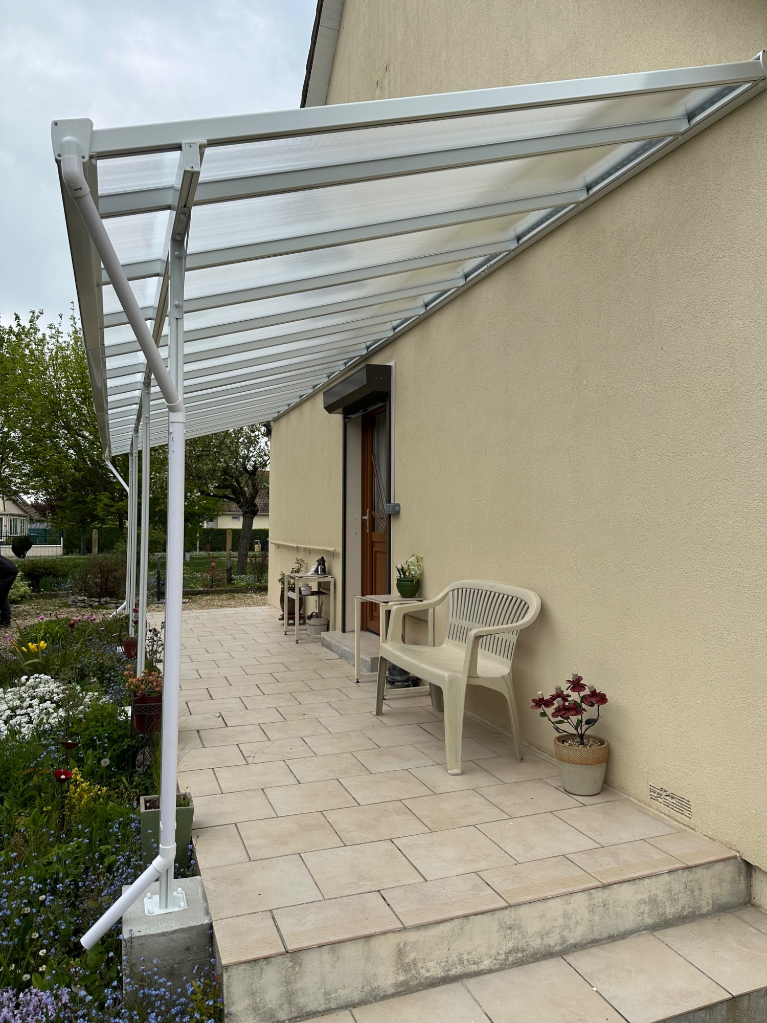 Auvent transparent en verre au-dessus d'une terrasse en pierre, avec une chaise et des plantes.