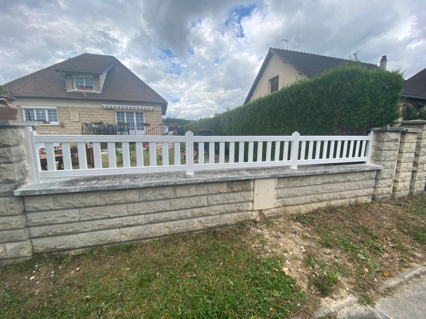 Clôture blanche en bois sur un muret en pierres, maison en arrière-plan.