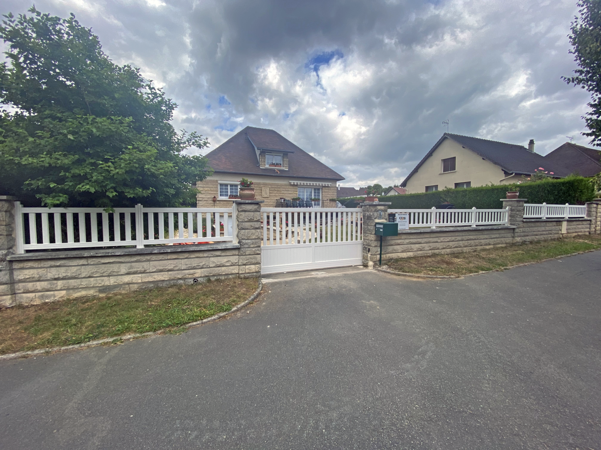 Maison avec clôture blanche et portail, située à un coin de rue sous un ciel nuageux.