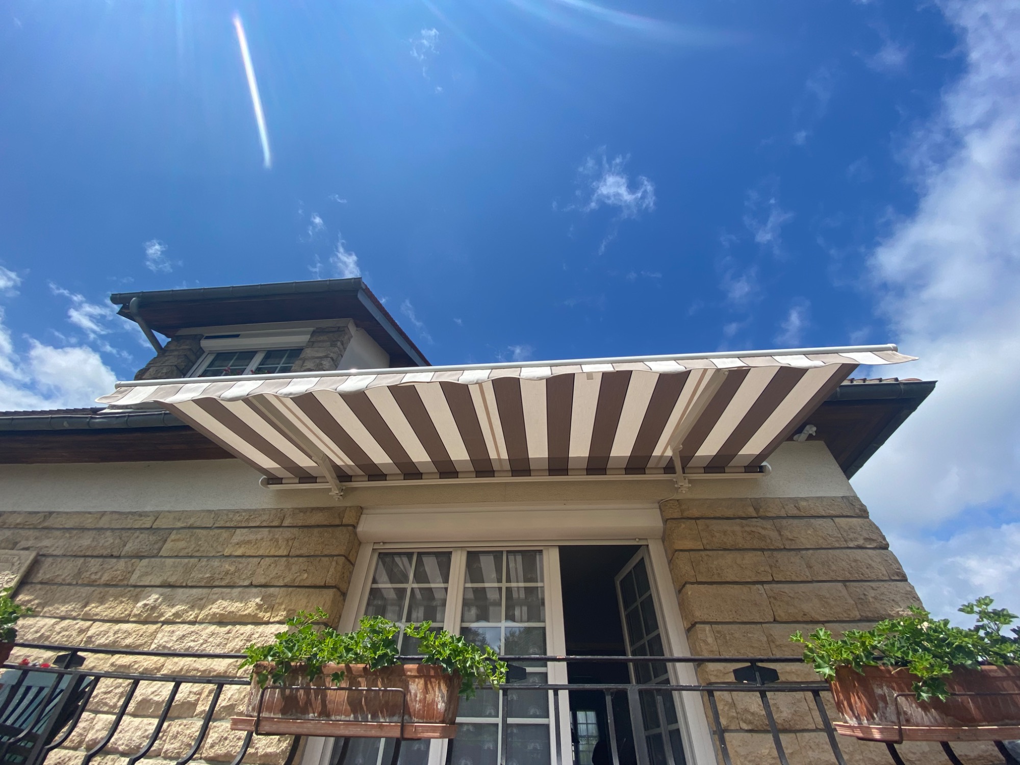 Maison avec auvent rayé, fleurs en pot et ciel bleu parsemé de nuages.
