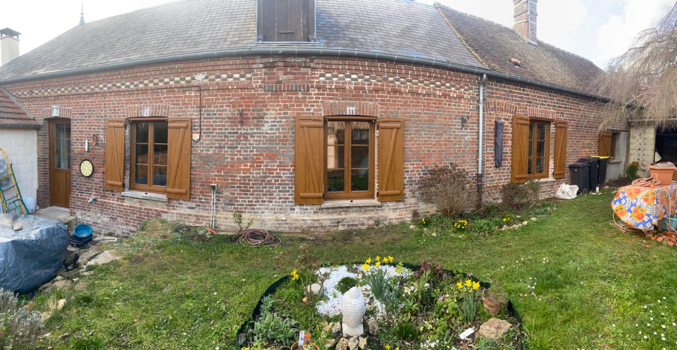 Vue panoramique d'une maison en briques avec des volets en bois et un jardin fleuri.