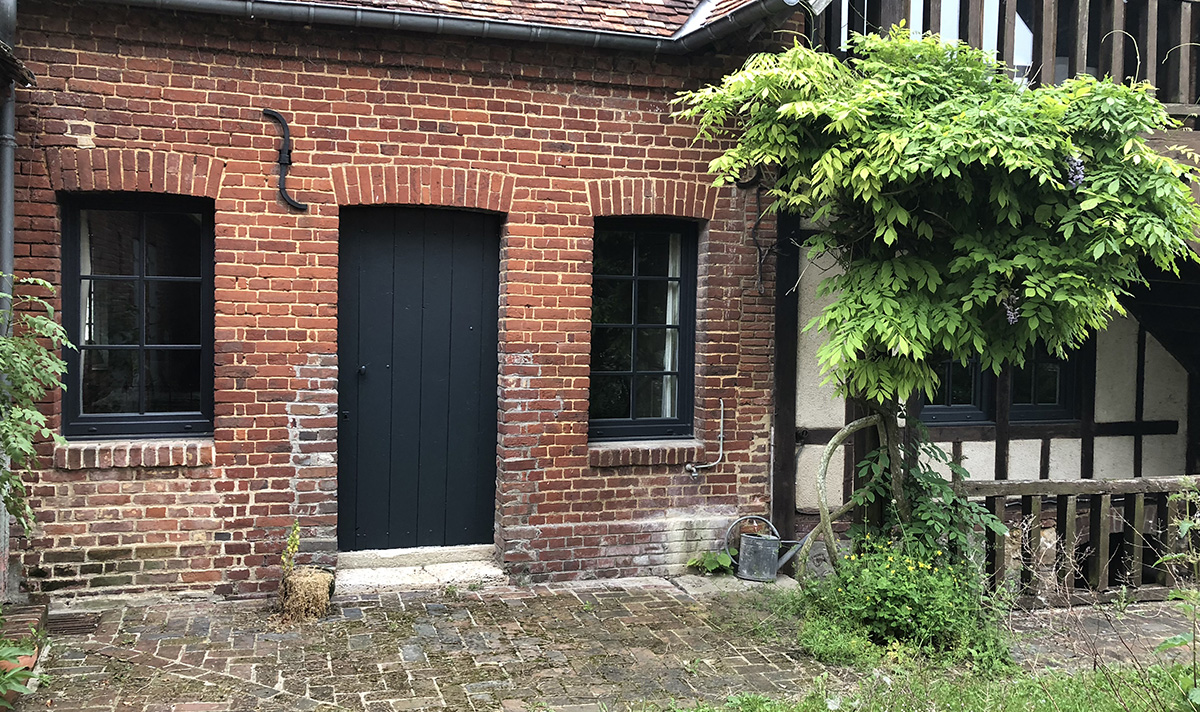Façade en briques rouges avec une porte noire et des fenêtres, entourée de verdure.