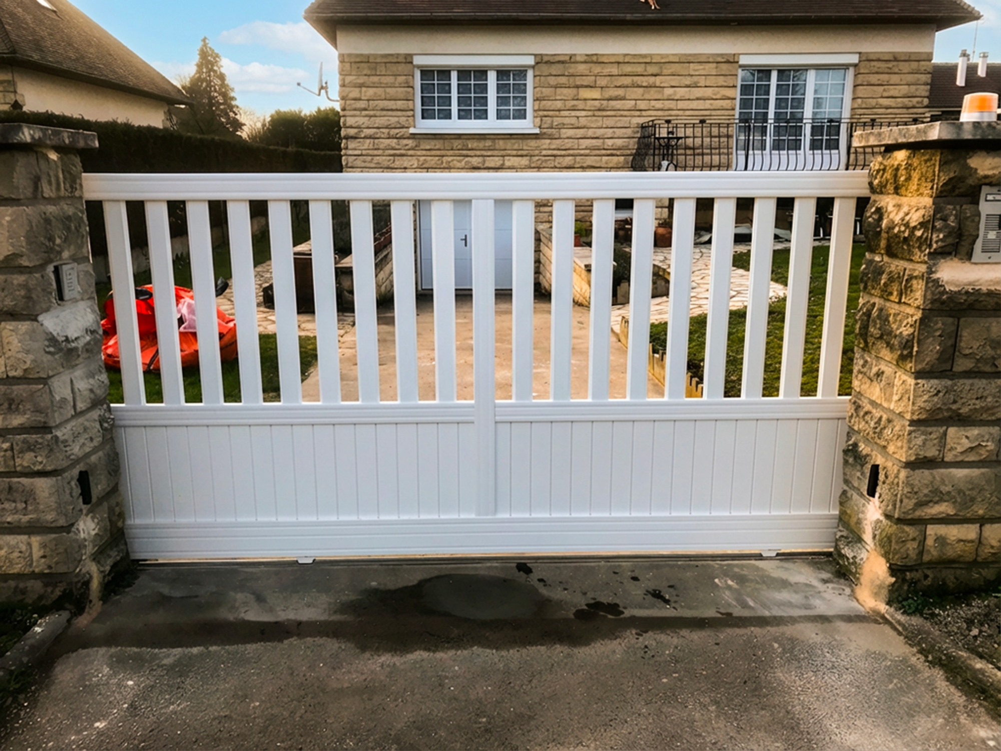 Portail en bois blanc avec des piliers en pierre, en arrière-plan une maison et un jardin.