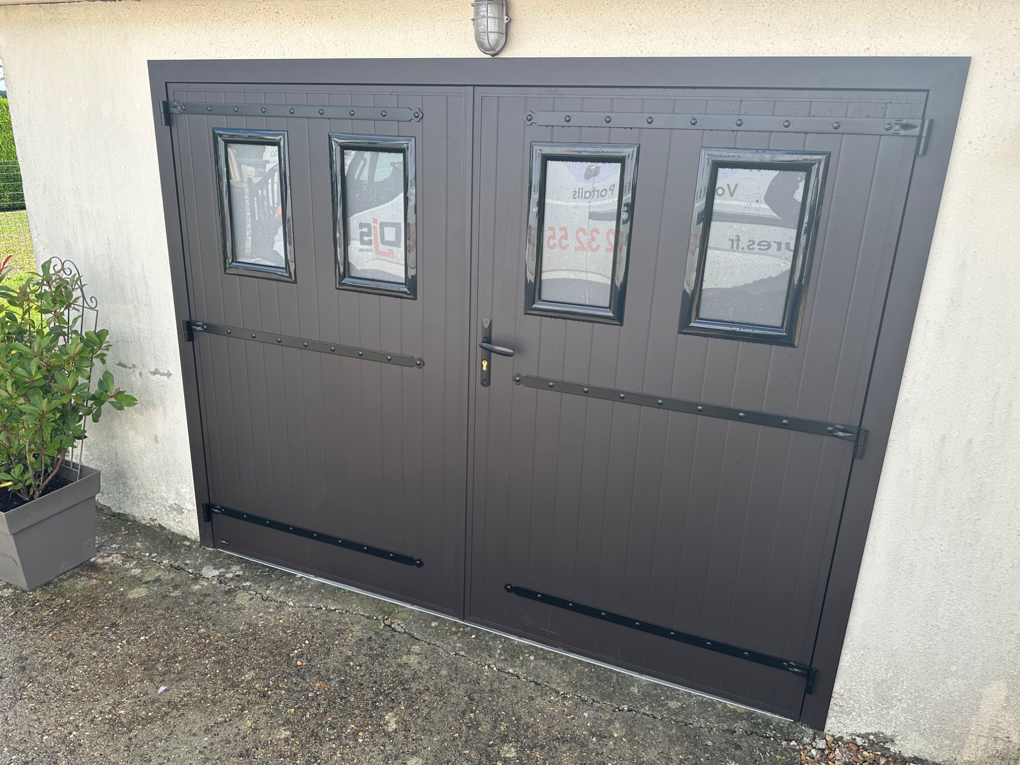 Porte de garage en métal gris avec fenêtres à petits carreaux. Plante en pot à gauche.