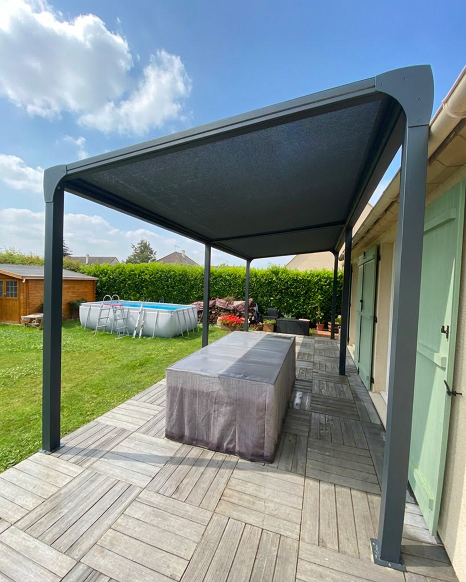 Auvent en métal noir au-dessus d'une table grise dans un jardin ensoleillé.