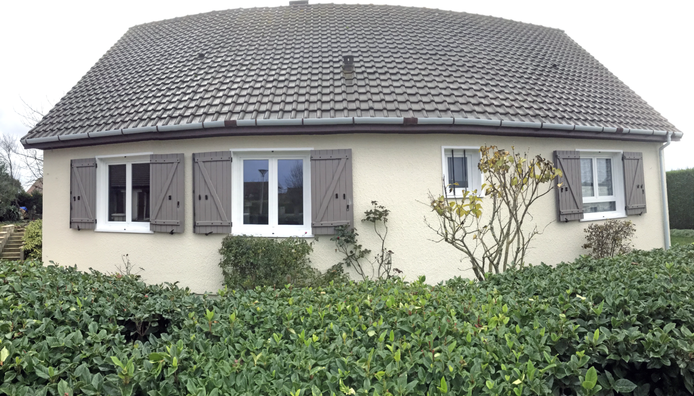 Maison de plain-pied avec fenêtres à volets, entourée de buissons.