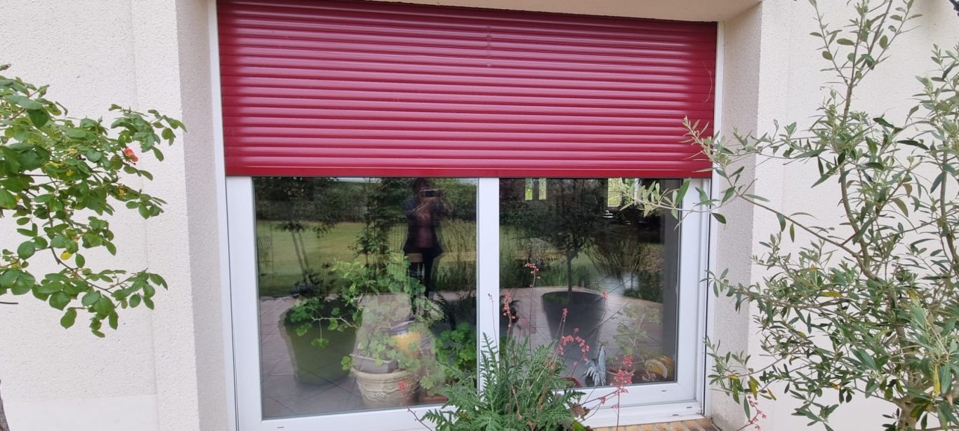 Volet roulant rouge devant une fenêtre avec des plantes à l'intérieur.