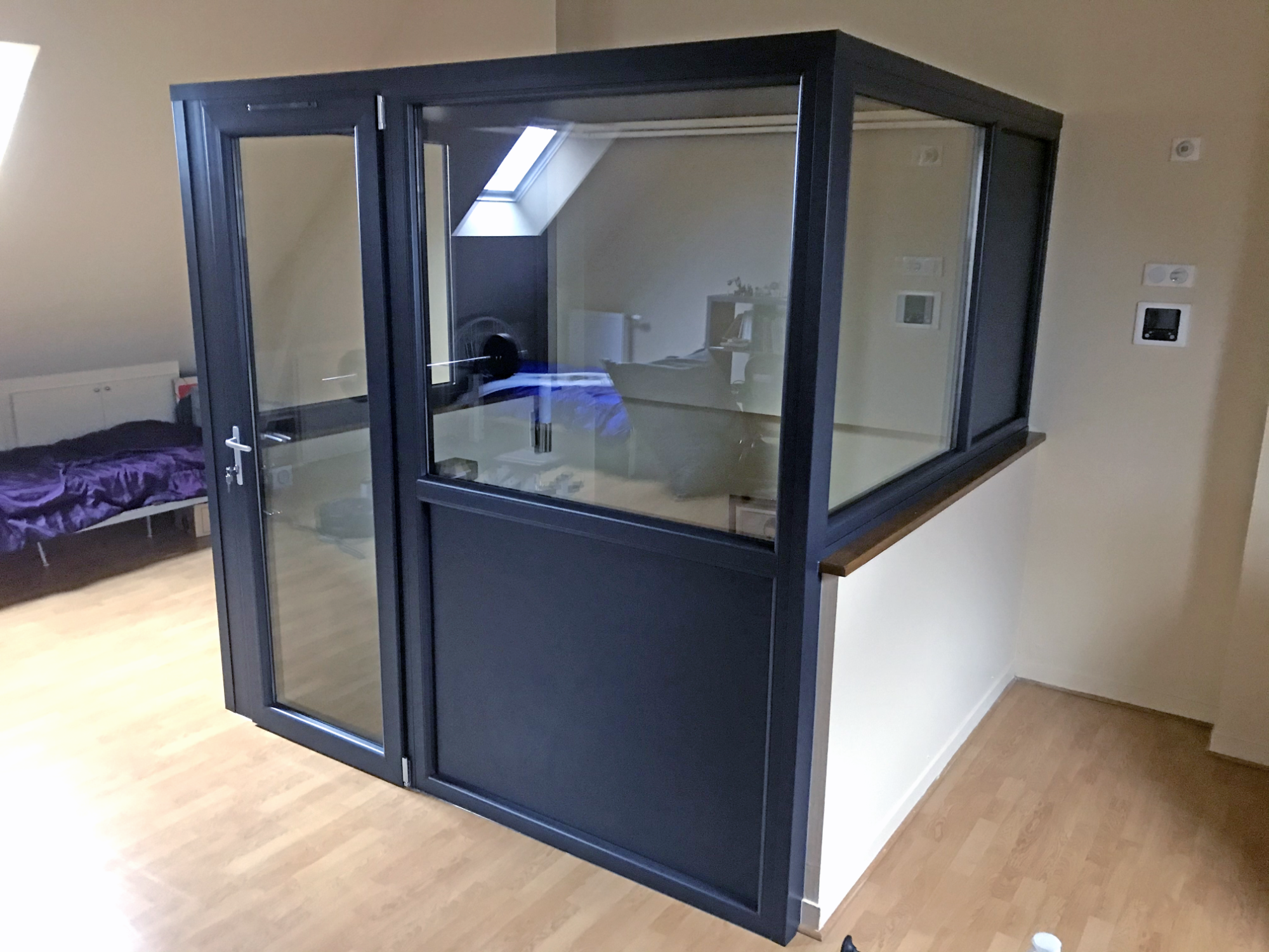 Cabine en verre avec des murs en métal, située dans une pièce au sol en bois.