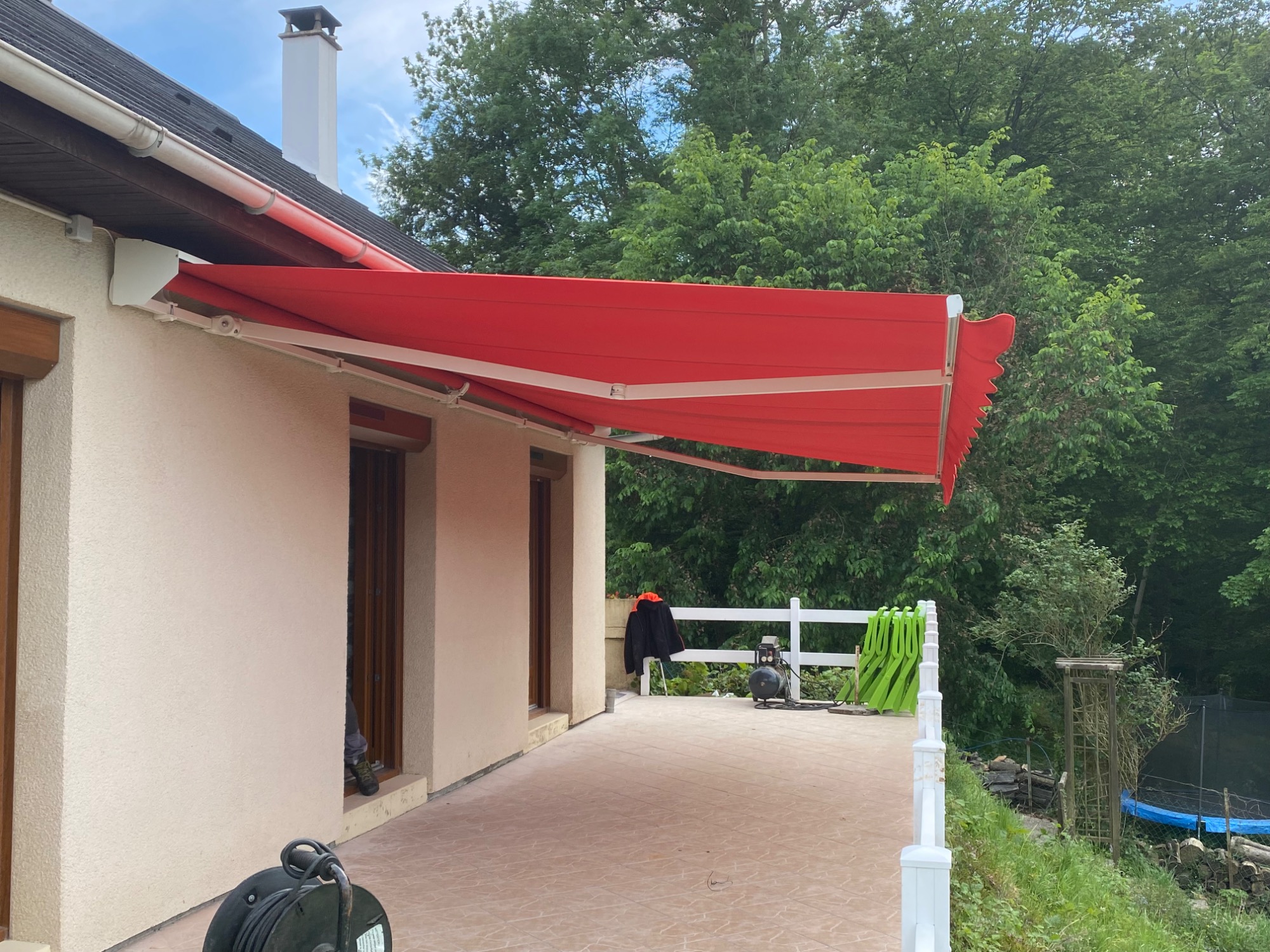 Auvent rouge sur une terrasse d'une maison, entourée d'arbres et d'un plan d'eau.