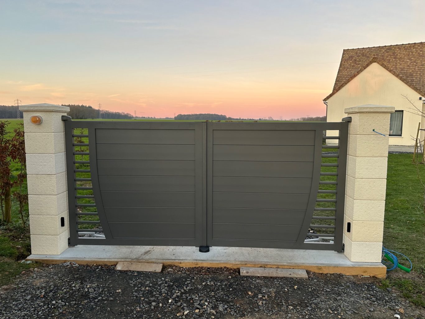 Portail en métal gris, avec des colonnes en pierre, devant un champ au crépuscule.