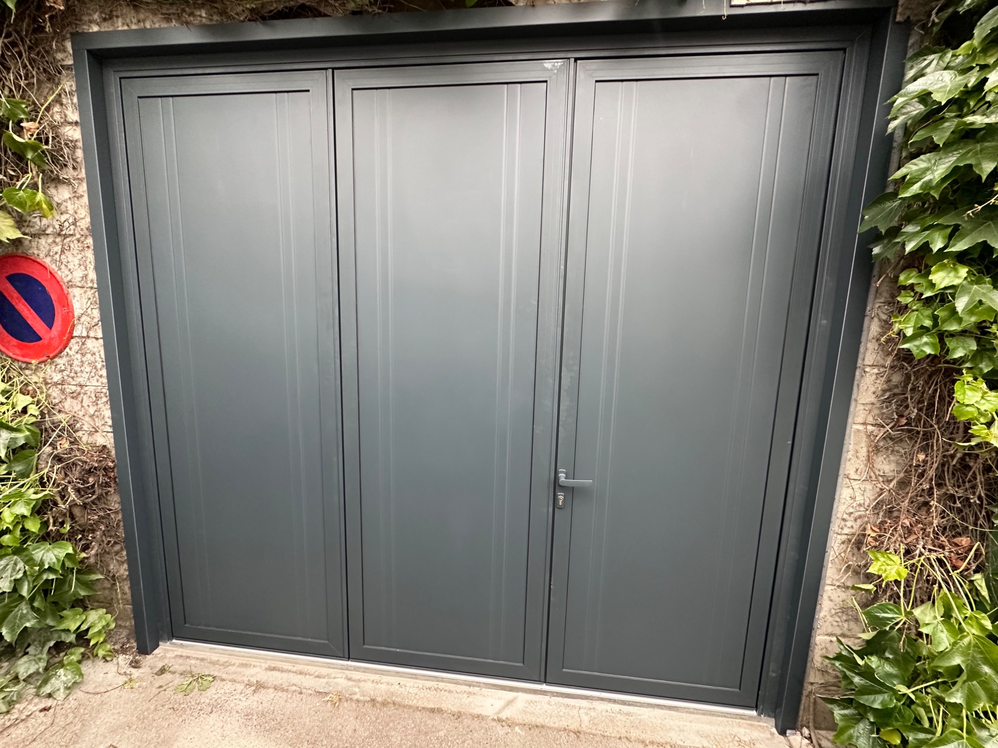 Porte de garage grise à trois sections, entourée de verdure.