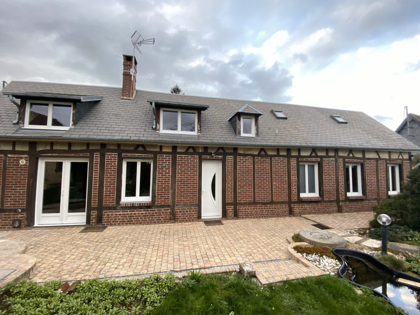 Maison en briques avec une toiture grise et un jardin avant bien entretenu.